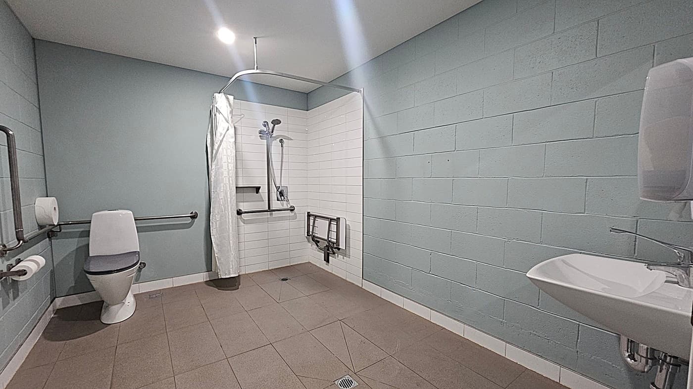A spacious, accessible bathroom with a beige tiled floor and white brick walls. On the left, there's a toilet with grab bars and a toilet roll holder. In the centre, a shower area with a curtain, handheld showerhead, and a foldable seat. On the right, a white sink with a lever tap and a paper towel dispenser. The ceiling is plain with a recessed light.