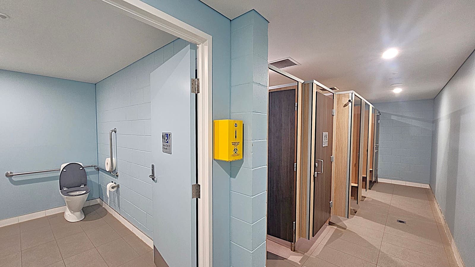 A public restroom with light blue walls and beige tiled flooring. On the left, there's an accessible toilet with grab bars and a toilet paper holder. A yellow sharps disposal box is mounted on the wall near the entrance. On the right, there are several wooden cubicle doors, each with a handle and a sign. The ceiling is white with recessed lighting.