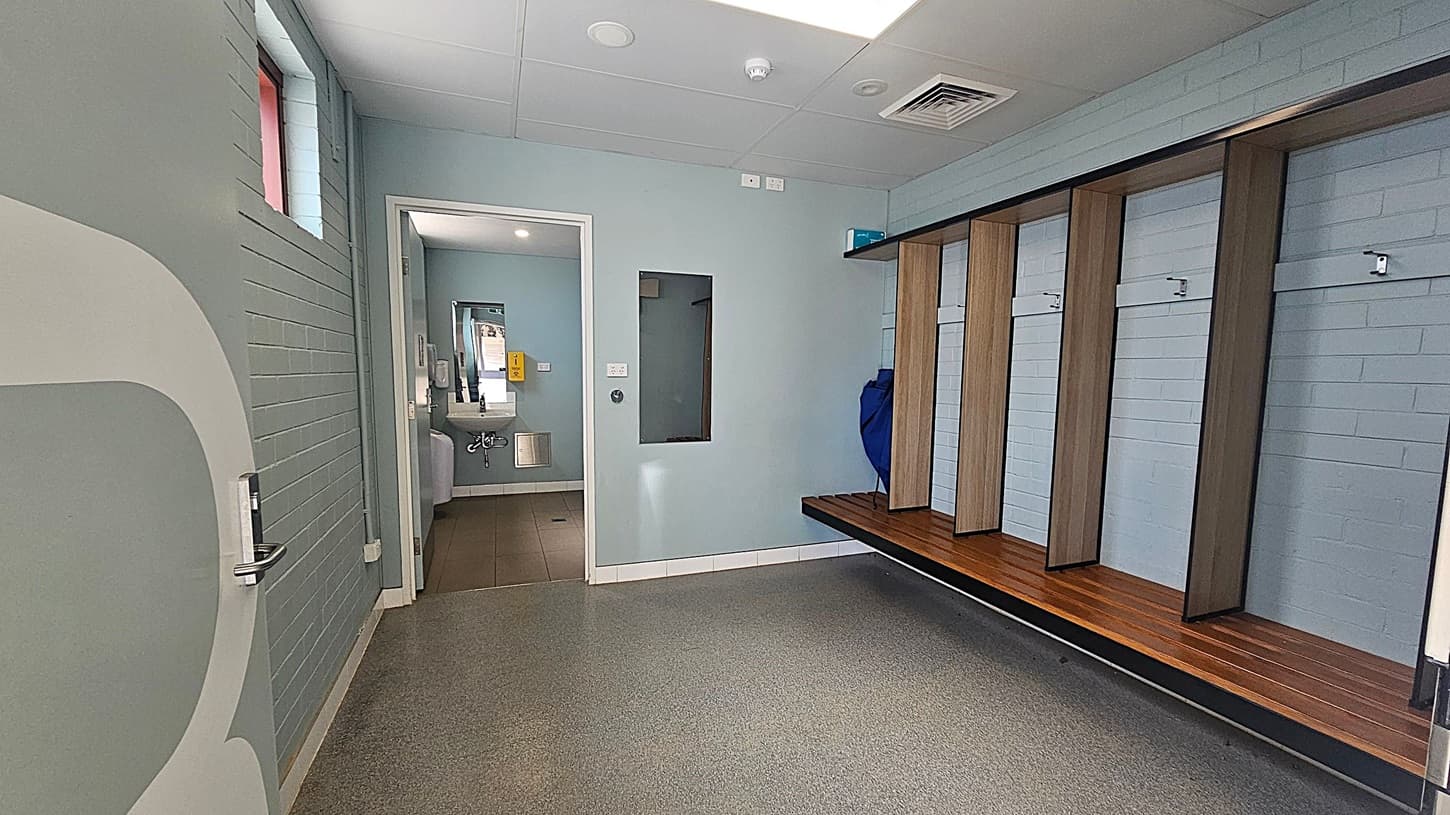 A changing room with a grey and green colour scheme. On the right, there are wooden benches with open cubbies above them. The floor is speckled grey and green. On the left, a partially open door leads to a corridor with a small window. In the centre, a doorway opens to a bathroom area with a visible sink and mirror. The ceiling is white with recessed lighting and ventilation panels.