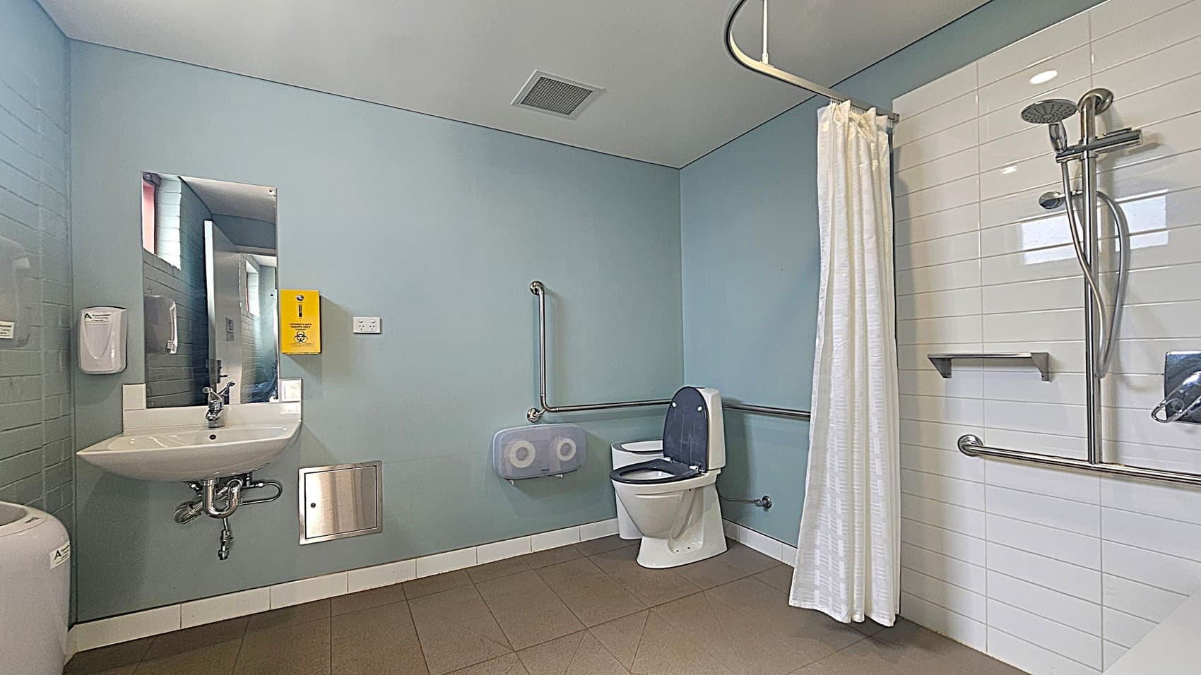 A spacious, accessible bathroom with light blue walls and a white ceiling. On the left, there's a sink with a mirror above it, a soap dispenser, and a yellow disposal box. The centre features a toilet with grab bars and a toilet paper dispenser. On the right, a shower area with a white curtain, handheld showerhead, and additional grab bars. The floor is tiled in a light brown colour.