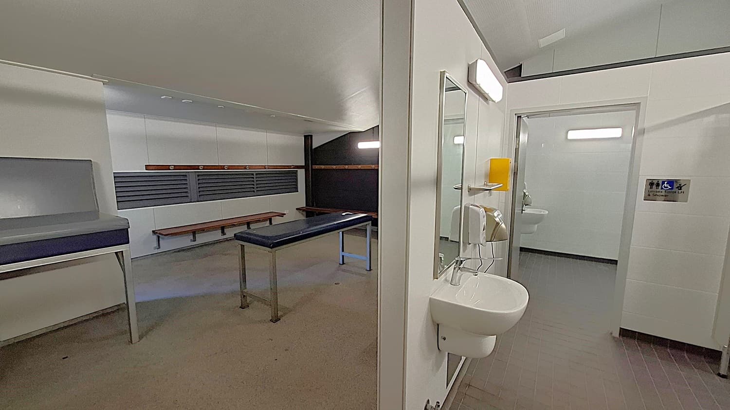 A room with a grey examination table in the centre, surrounded by benches along the walls. On the left, there's a cushioned bench. A sink with a mirror and soap dispenser is mounted on a dividing wall. To the right, an open doorway leads to a bathroom with a visible sink and a sign indicating accessibility. The floor is tiled in dark grey, and the ceiling is light-coloured.
