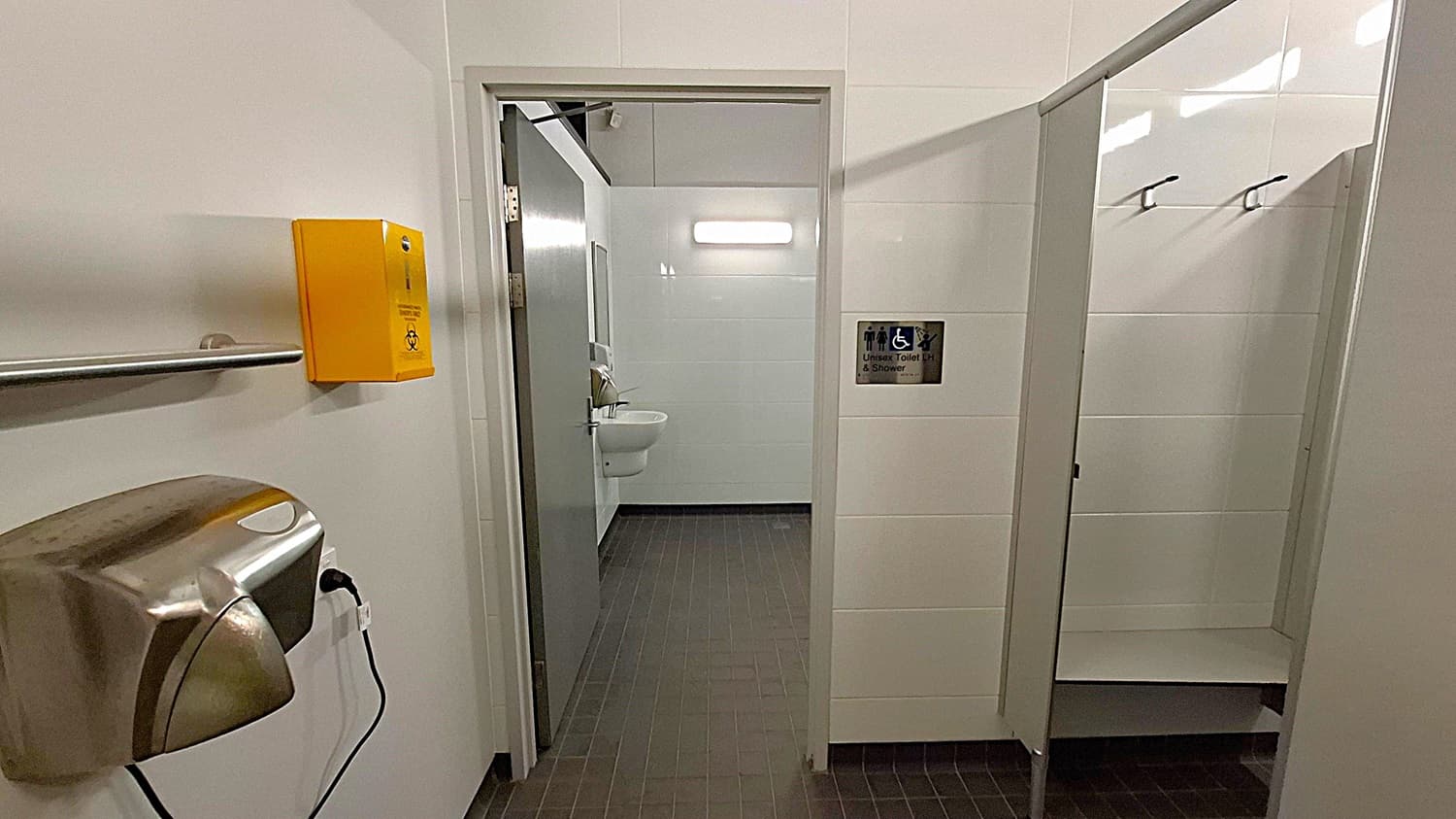 A public restroom with a grey tiled floor and light-coloured walls. On the left, there's a stainless steel hand dryer and a yellow biohazard disposal box mounted on the wall. In the centre, an open door leads to a room with a sink and a mirror, illuminated by a wall light. On the right, a sign indicates an accessible unisex toilet and shower. There are hooks on the wall inside the shower area.