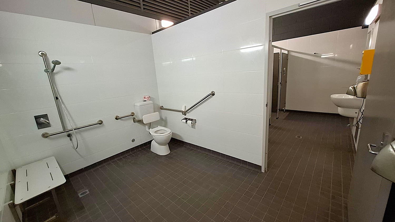 A public accessible bathroom with white tiled walls and dark tiled floor. On the left, there's a shower area with a handheld showerhead and a foldable bench. In the centre, a toilet with grab bars on both sides and a toilet paper holder. The door on the right leads to another area with a sink and hand dryer. The ceiling has a grid-like design with fluorescent lighting.