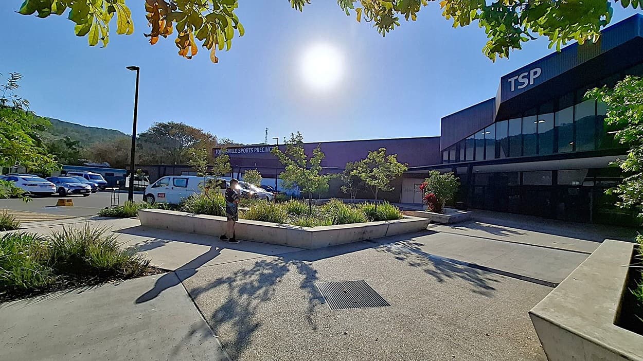 A modern building with "TSP" on the facade is on the left, featuring large glass windows. In front, a white van is parked, and a hand truck is on the pavement. The centre has a landscaped area with plants and a pathway leading to another structure with a slanted roof. On the right, there are more trees and a person walking. The ground has yellow-striped markings, and there are tall street lamps.