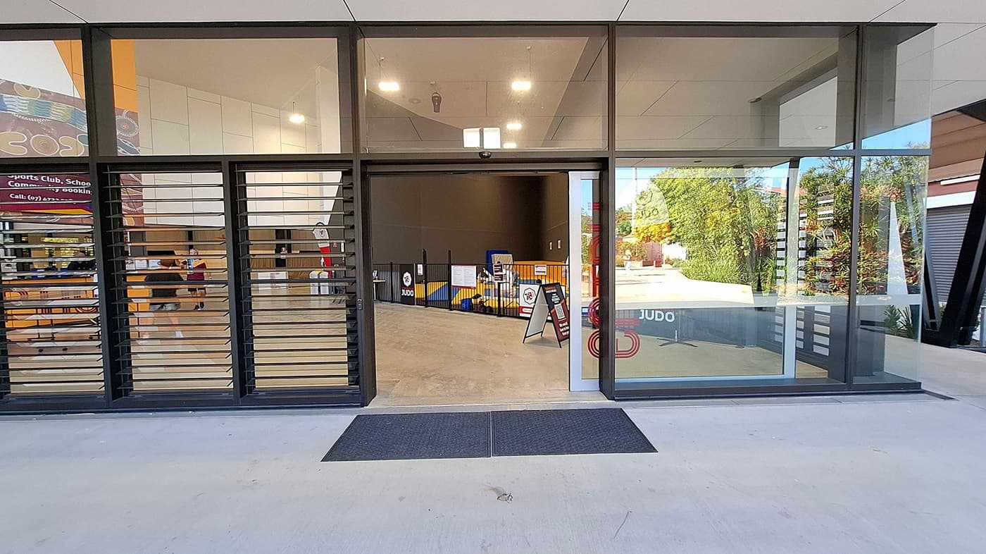 Glass entrance with sliding doors and louvred windows on the left. Inside, a spacious area with a light-coloured floor and high ceiling. A judo mat area is visible, enclosed by a low fence with signs. A-frame signs are placed near the entrance. On the right, large windows show greenery outside. The ceiling has recessed lighting, and there are two black mats on the ground outside the entrance.