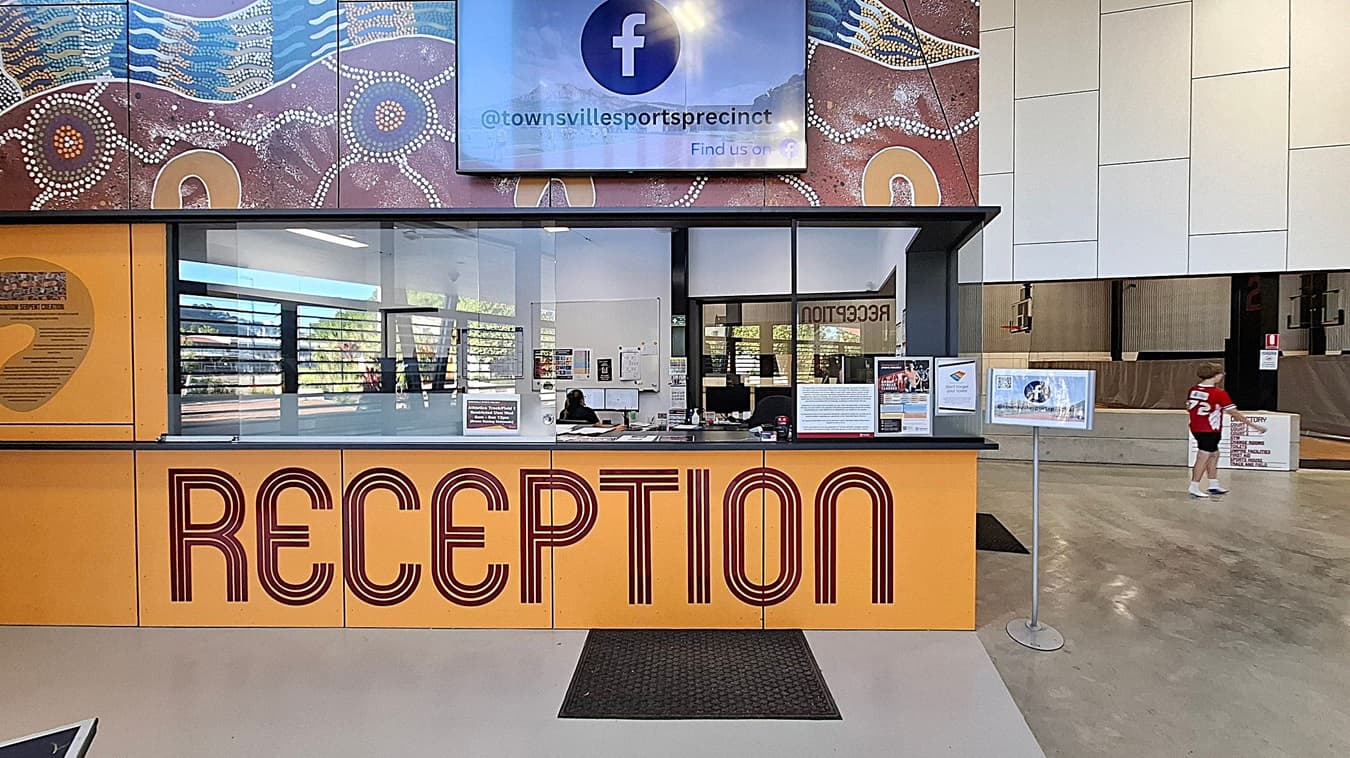 Reception area with a large yellow desk featuring the word "RECEPTION" in bold red letters. Behind the desk, there's a glass partition with a person seated at a computer. Above, a digital screen displays a Facebook handle. The wall has an Aboriginal art design with earthy tones and dot patterns. To the right, a child in a red shirt walks past a sign. The floor is a mix of grey and green, and the ceiling is not visible.
