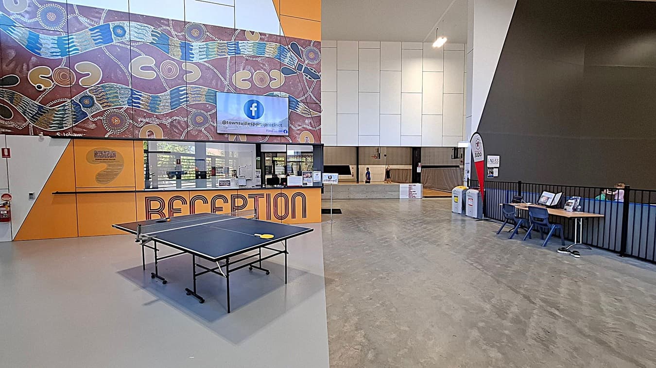 Reception area with a large yellow desk featuring the word "RECEPTION" in bold red letters. Behind the desk, there's a glass partition with a person seated at a computer. Above, a digital screen displays a Facebook handle. The wall has an Aboriginal art design with earthy tones and dot patterns. To the right, a child in a red shirt walks past a sign. The floor is a mix of grey and green, and the ceiling is not visible.