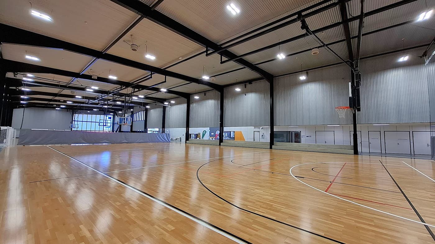 A spacious indoor sports hall with a polished wooden floor marked for basketball. On the right, a basketball hoop is mounted on a black metal frame. The walls are grey with vertical panelling, and there are several doors and windows along the right side. The ceiling is high with exposed beams and bright overhead lights. In the background, a large window allows natural light to enter, and a grey divider partially separates the space.