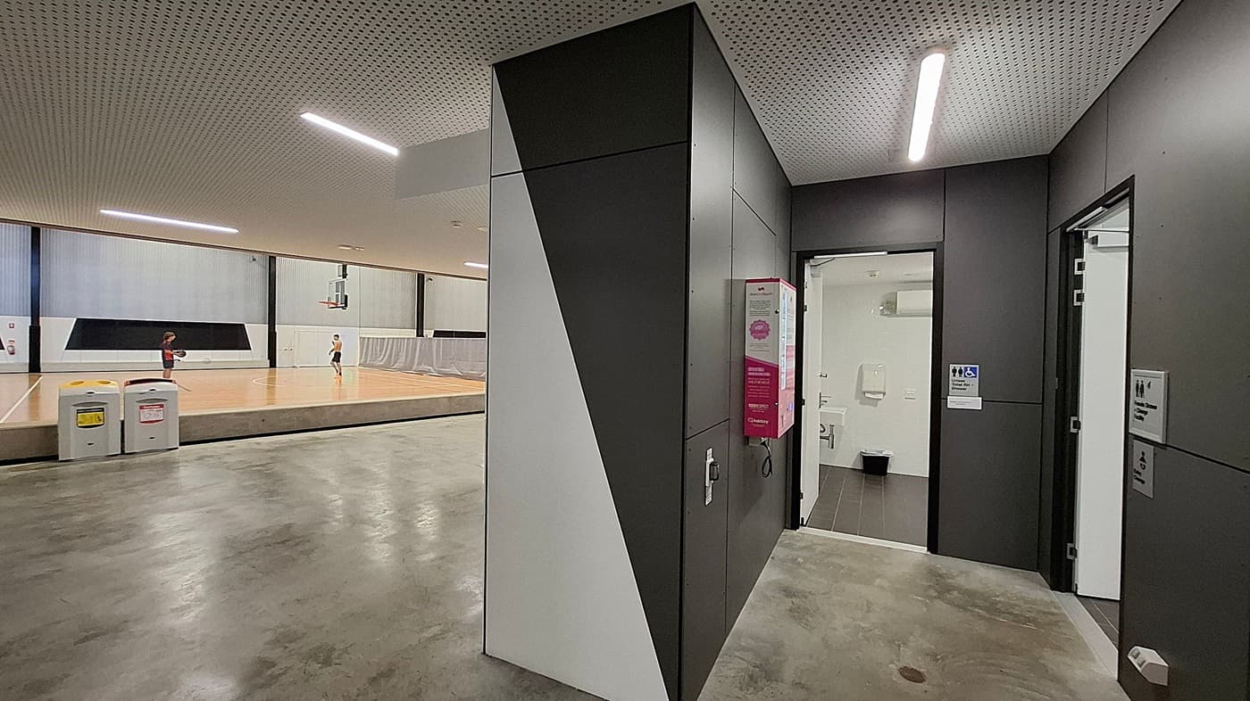 On the left, a basketball court with two people playing. The court has a wooden floor and a basketball hoop. In the foreground, there are two waste bins. In the middle, a black and white geometric wall structure. On the right, a corridor with a door leading to a restroom, marked with a sign for accessibility. The ceiling is perforated with linear lights, and the floor is a polished concrete surface.