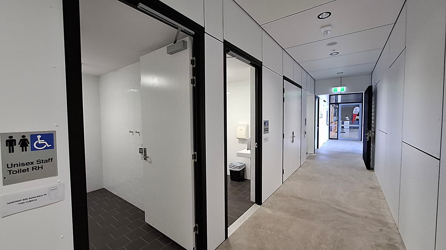 A modern building entrance with a covered walkway. On the left, there are symbols indicating accessible facilities, including a wheelchair and baby changing area. A glass door leads inside, with a fire hose reel and extinguisher nearby. The right side features large windows with horizontal slats. The ceiling is white with recessed lighting, and the floor is a smooth concrete surface. In the background, there are plants and a closed roller door.