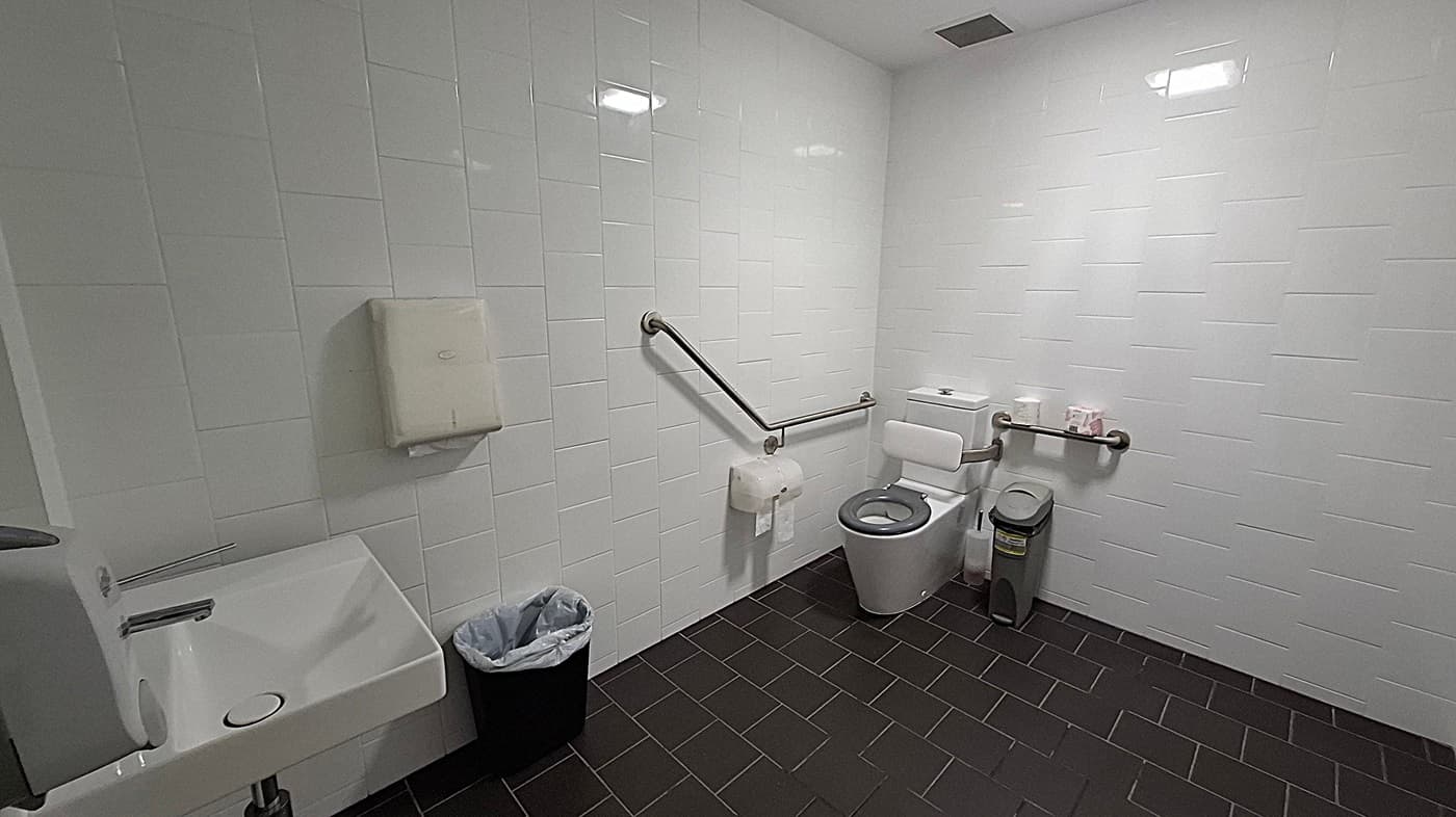 A corridor with white walls and a light-coloured floor. On the left, there are several open doors leading to unisex staff toilets, each with a sign indicating accessibility features. Inside the toilets, there are visible sinks and bins. The ceiling has recessed lighting. At the end of the corridor, there is a glass door leading to another area, with a sign visible through the glass.