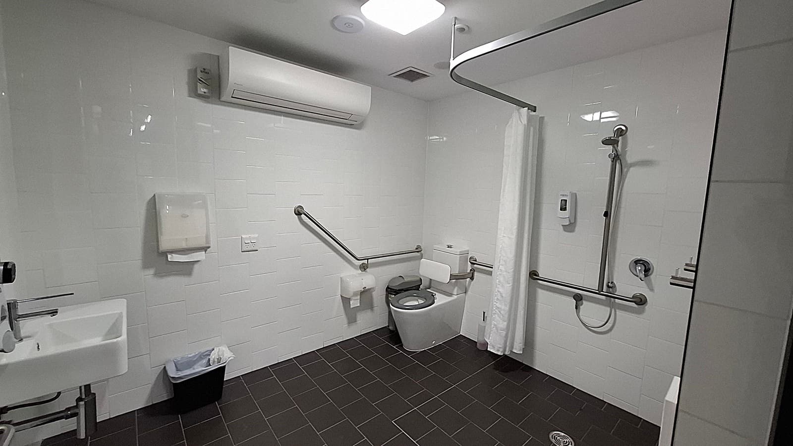 A public restroom with white tiled walls and a dark tiled floor. On the left, there is a white sink with a soap dispenser and a bin underneath. A green paper towel dispenser is mounted on the wall. In the centre, a toilet with support rails on both sides is visible. To the right of the toilet, there is a small bin and some toilet paper rolls on a shelf. The ceiling is plain with a light fixture.