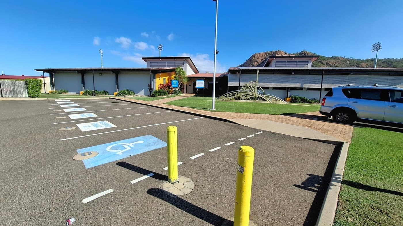 A parking area with marked spaces, including a blue disabled parking spot in the foreground. Two yellow bollards are near the entrance. On the right, a silver car is parked. In the background, there are modern buildings with a yellow entrance and a metal sculpture on a grassy area. A mountain with floodlights is visible in the distance. The ground is paved, and there are small plants along the building's edge.