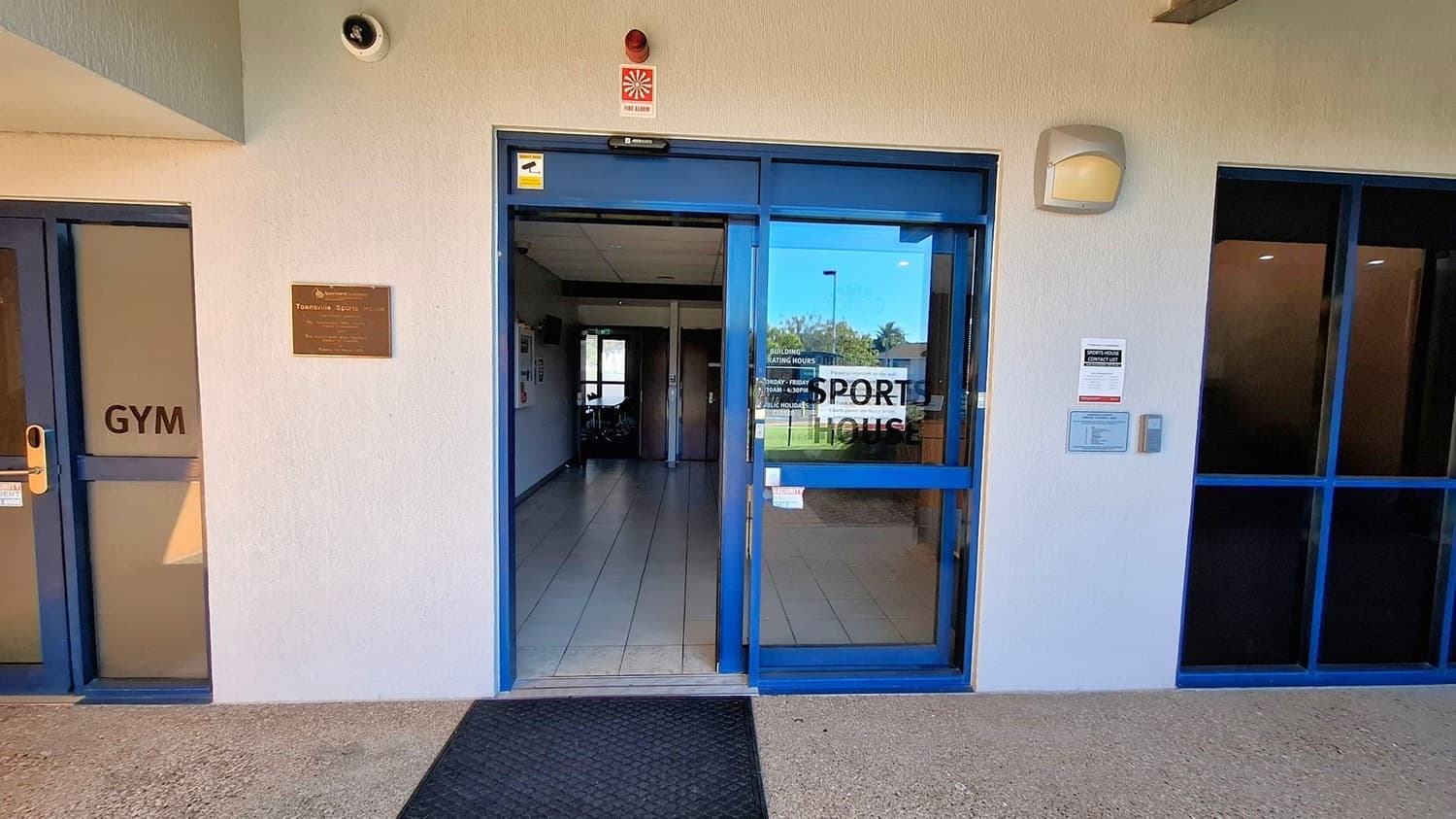 Entrance to a building with a blue-framed glass door in the centre, labelled "SPORTS HOUSE". To the left, a door marked "GYM" and a plaque reading "Townsville Sports House". Above the entrance, a fire alarm and security camera are visible. On the right, a notice board and intercom system are mounted on the wall. The floor is tiled, and a black mat is placed at the entrance.