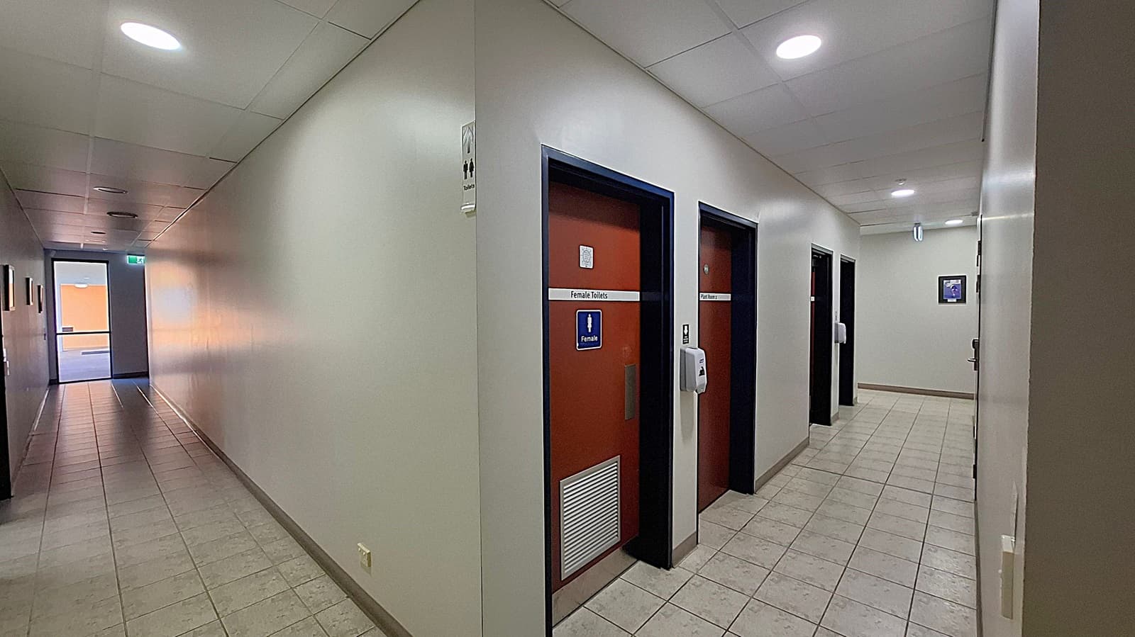 A corridor with tiled flooring and a ceiling with recessed lights. On the right, there are doors labelled "Female Toilets" and "Parent Room," each with a sign and a wall-mounted hand sanitiser dispenser. The left side of the corridor is a plain wall leading to a glass door at the end, allowing natural light to enter. A small framed picture is visible on the right wall near the end of the corridor.