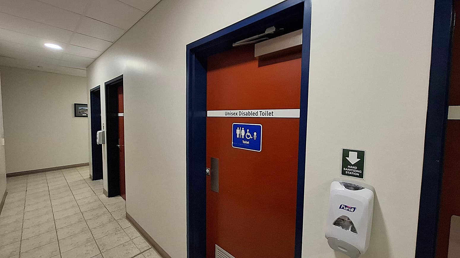 A unisex accessible toilet with a red door featuring a sign indicating accessibility. Inside, on the left, there is a toilet with grab bars and a roll of toilet paper on top of the cistern. A sanitary bin is nearby. In the centre, a paper towel dispenser is mounted above a bin. On the right, a sink with a mirror above it and a small shelf holding soap and reed diffusers. The floor is tiled in grey, and the walls are light-coloured.