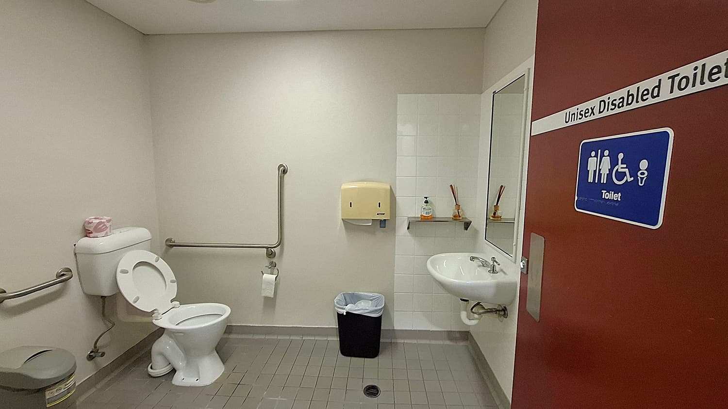 A unisex accessible toilet with a red door featuring a sign indicating accessibility. Inside, on the left, there is a toilet with grab bars and a roll of toilet paper on top of the cistern. A sanitary bin is nearby. In the centre, a paper towel dispenser is mounted above a bin. On the right, a sink with a mirror above it and a small shelf holding soap and reed diffusers. The floor is tiled in grey, and the walls are light-coloured.