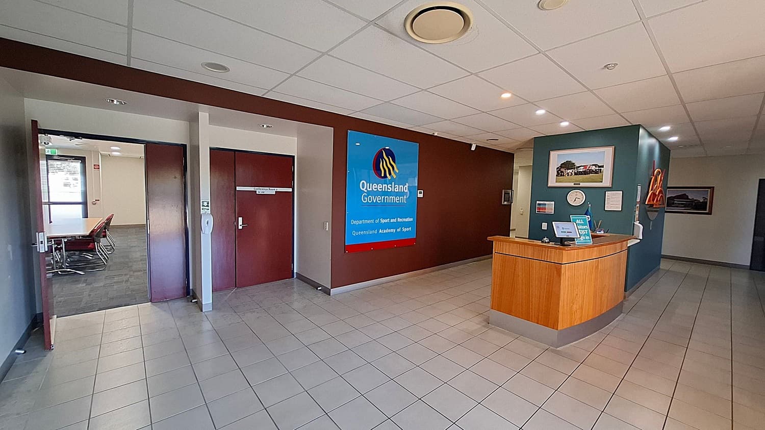 A reception area with a tiled floor and a suspended ceiling with lights. On the left, there are two wooden doors, one open, leading to a room with a table and chairs. In the centre, a large sign reads "Queensland Government, Department of Sport and Recreation, Queensland Academy of Sport." On the right, a wooden reception desk with a computer and a sign. Behind the desk, a wall with a clock, a framed picture, and some orange safety equipment.