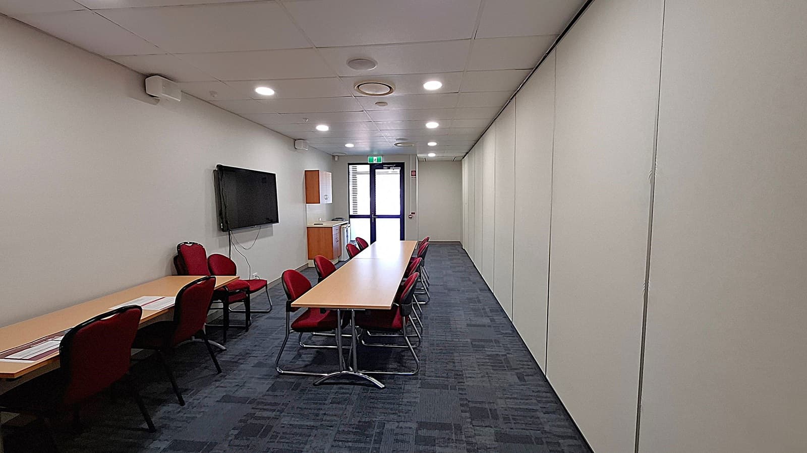 A conference room with a long wooden table in the centre, surrounded by red chairs. On the left wall, there's a large flat-screen TV mounted, and a small wooden cabinet beneath it. The right wall is plain and white. At the far end, there's a glass door with an exit sign above it. The floor is carpeted in a dark pattern, and the ceiling is fitted with recessed lights.