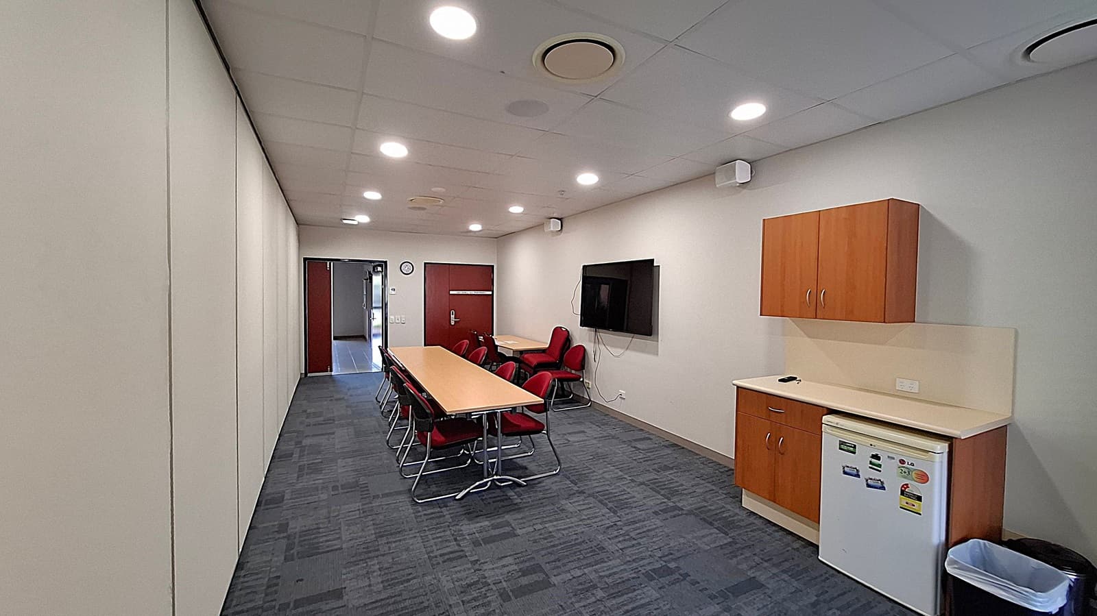 A conference room with a long table and red chairs in the centre. On the right, there's a wall-mounted TV and a kitchenette with wooden cabinets, a small fridge, and a bin. The left wall is plain, and the floor is carpeted in a dark pattern. The ceiling has recessed lighting and air vents. At the far end, there's a door and a clock on the wall.