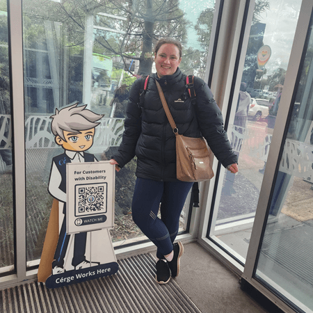A woman in a black jacket stands beside a Cérge cartoon cutout with a QR sign.