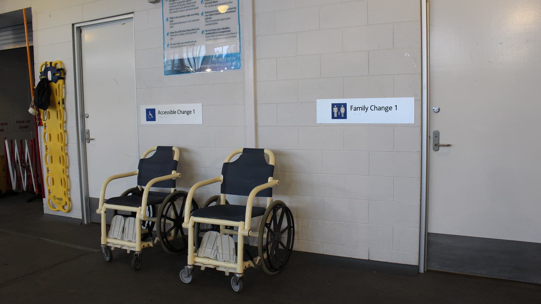 There are two aquatic wheelchairs situated between the entrances to the ‘Accessible Change ‘1 and the ‘Family Change 1’ and there is signage above these wheelchairs providing instructions to visitors. To the left there is a yellow, spinal board hanging on the wall.
