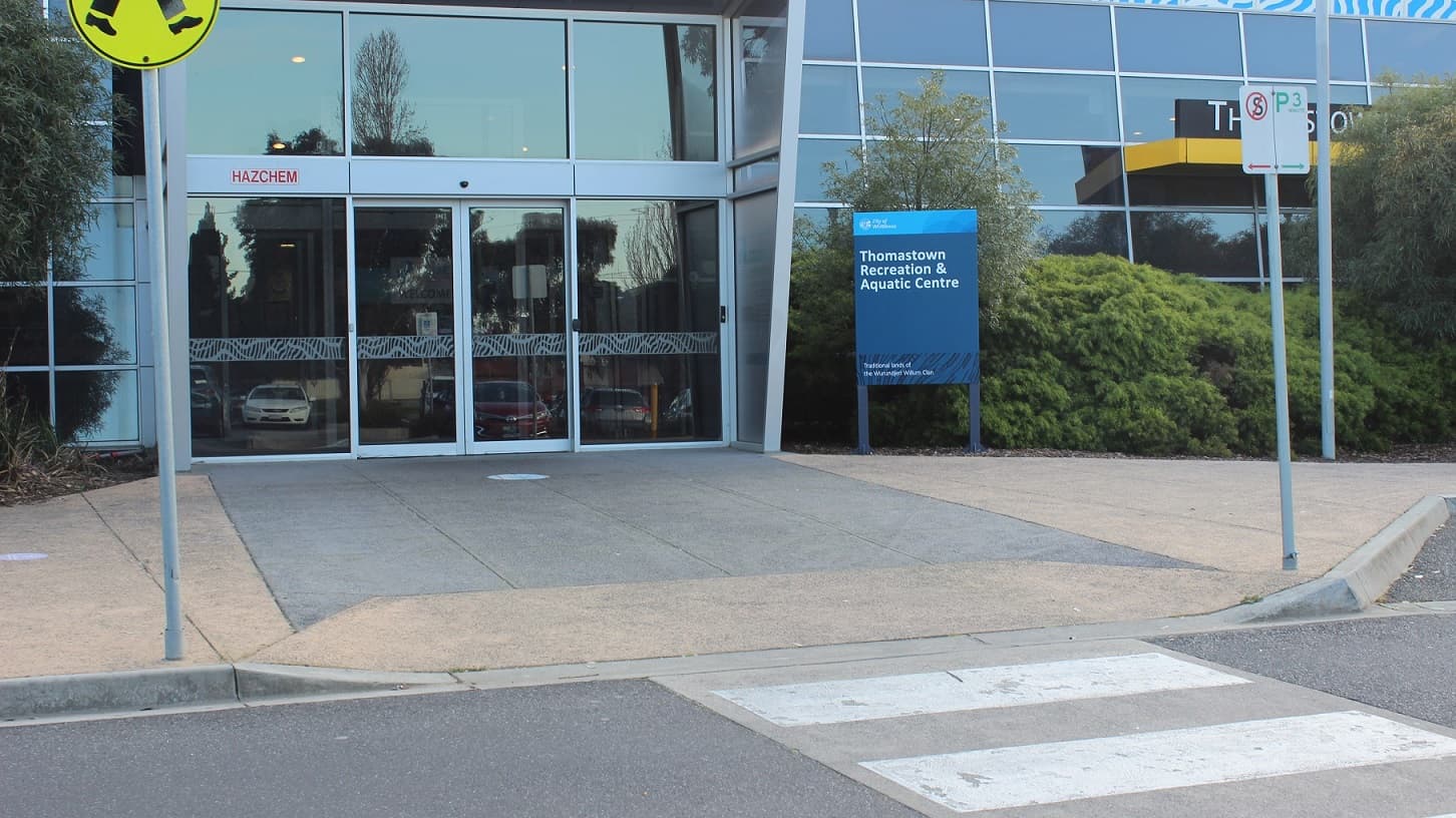 This image is a closer view of the previous image and is focusing in on the blue signboard with white writing welcoming visitors to the Thomastown Recreation & Aquatic Centre. Signage above the left hand side glass sliding door states “HAZCHEM”. Light poles and pedestrian signage is situated around the entrance