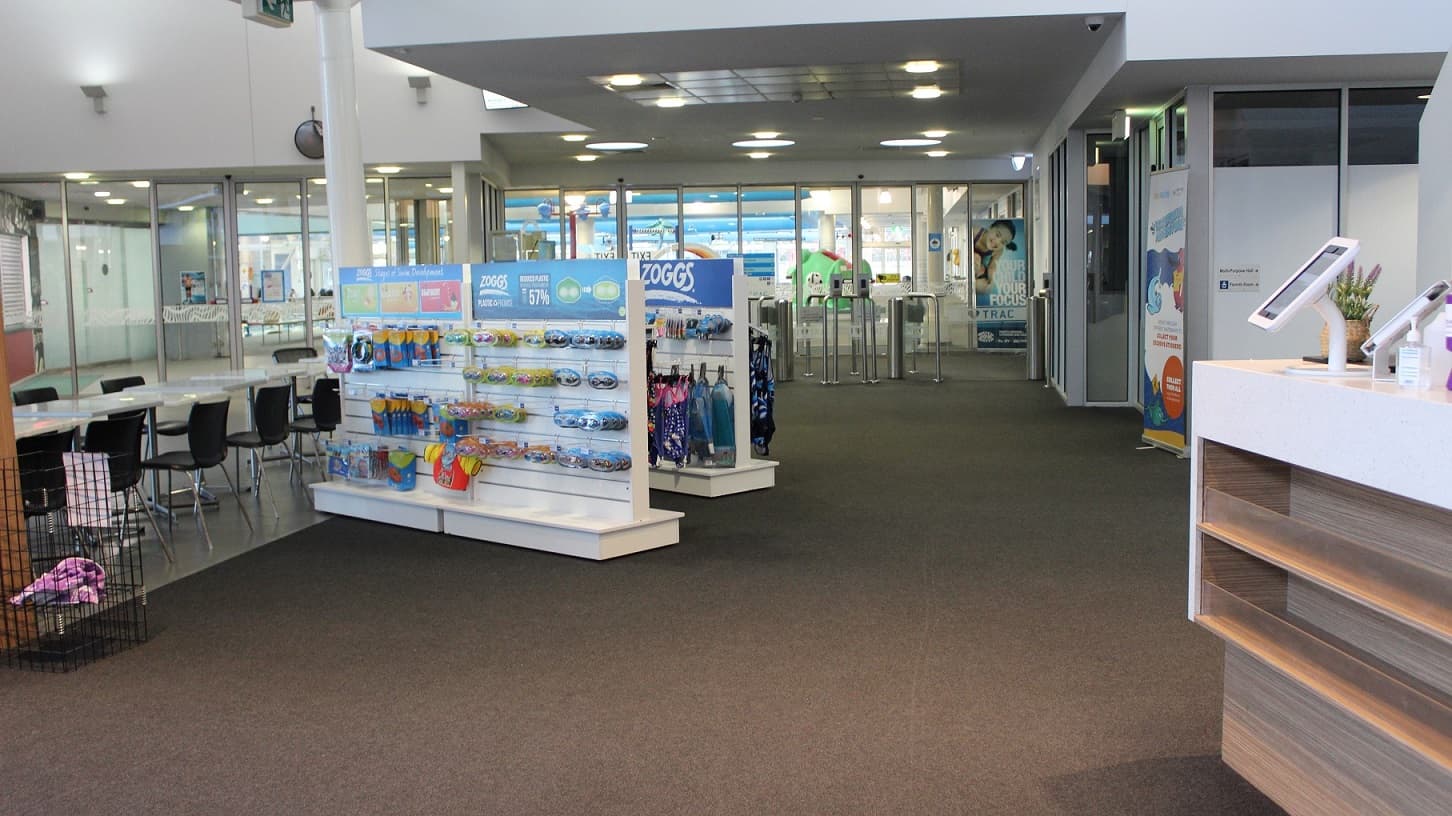 This image is a closer look of the previous image and features the two racks of swimming gear and equipment for sale. Further to the left are a range of white tables and black chairs. In the distance through the glass walls is the indoor aquatic centre.
