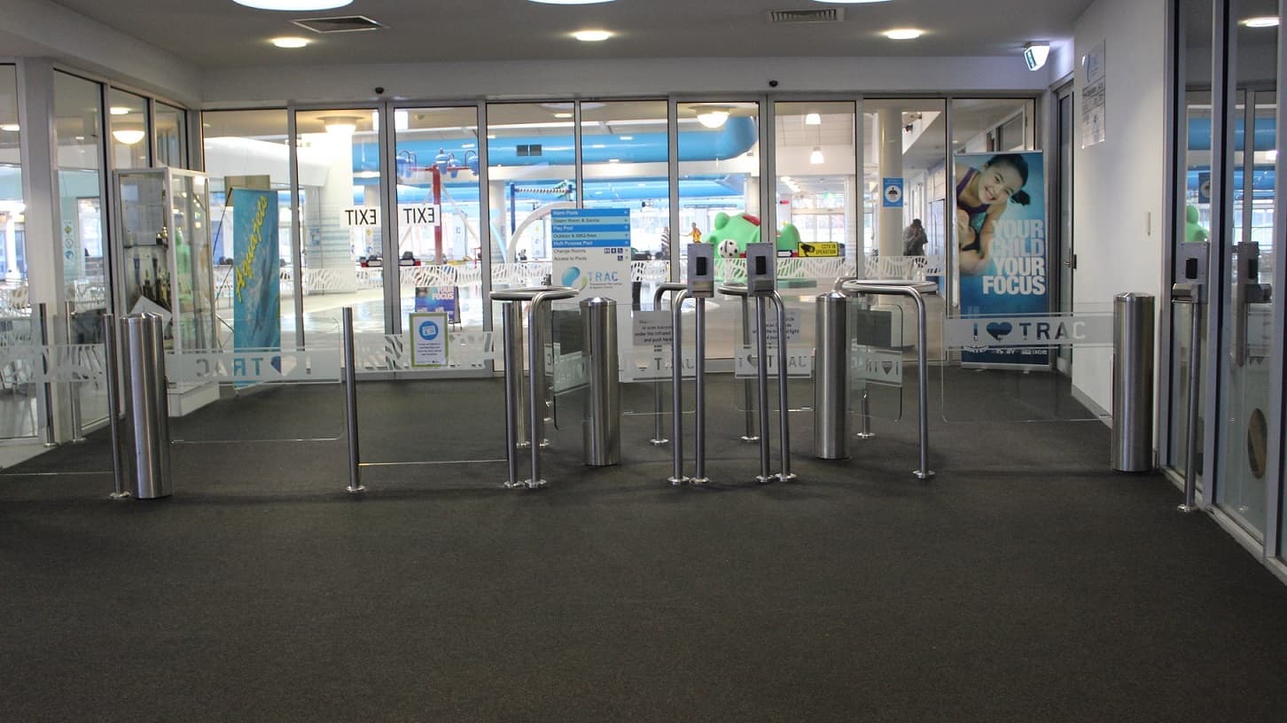 Directly ahead are the glass and steel turnstiles that lead into the indoor aquatic centre. To the left and to the right is wheelchair access through the turnstiles. The glass walls are covered with information and directional signage. “I love TRAC '' is written in white across the glass panels of the turnstiles. The children's leisure pool can be viewed in the distance.