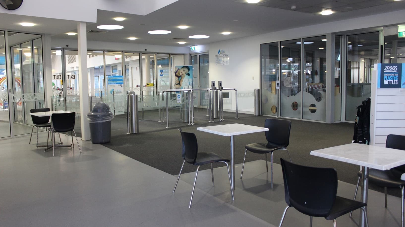 In this brightly lit seating area there are six black chairs and three white tables, with each table having two chairs placed at it. A rubbish bin leans on a white pillar by the exit turnstile gate to the left. A “Zoggs” promotional sign is on display on the wall by the right overlooking two sets of chairs and tables.