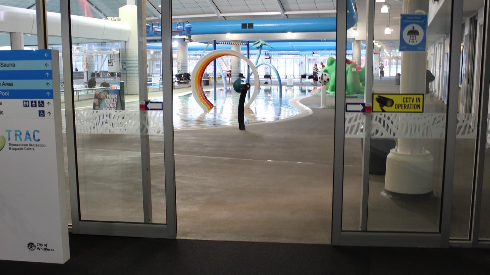 To the left of the automatic glass sliding door, there is a white and blue sign board providing directions to the various areas of the facility. Directly ahead is the children's water park that features a green frog slide to the right and in the centre of the water are round pipes with white, red, yellow and orange trim. The pool has a noticeboard at one end stating “Your Child, Your Focus”.