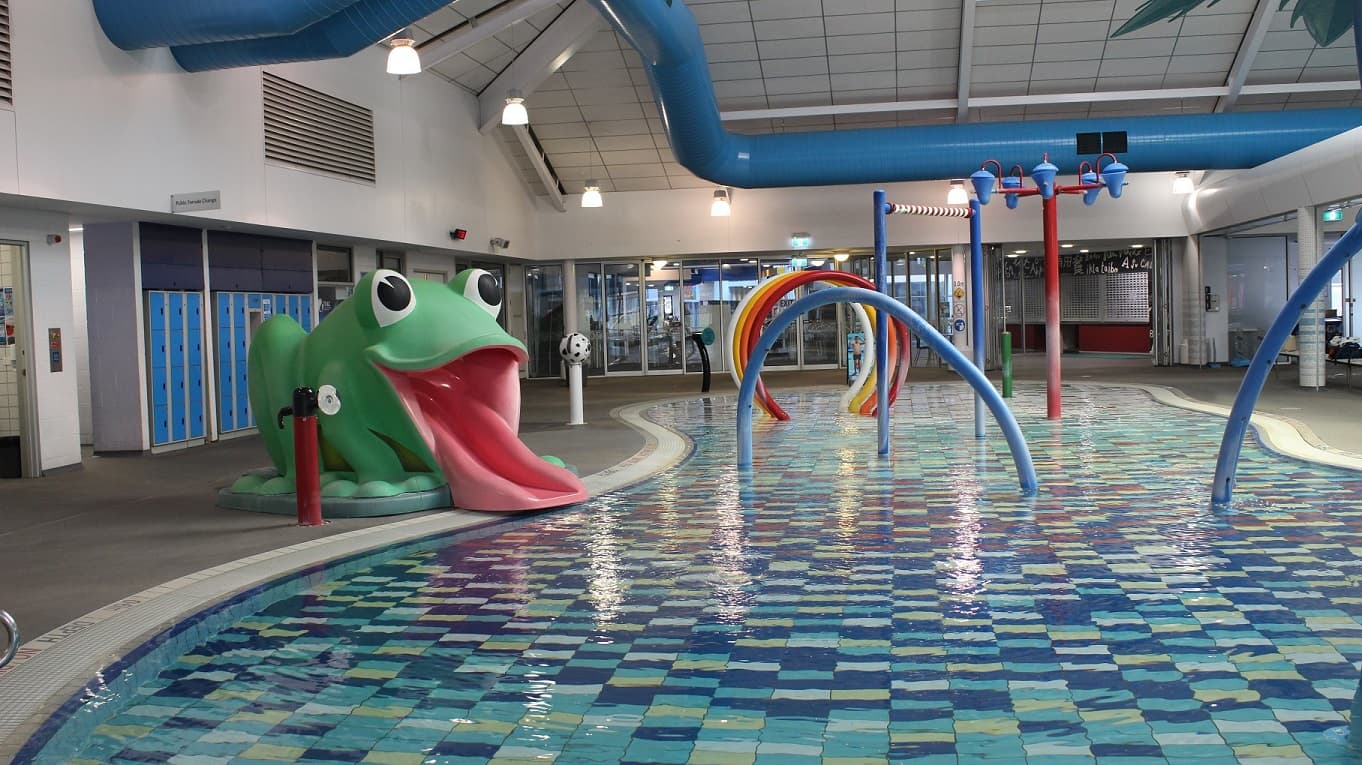 The children's leisure pool, tiled in blue, white and grey, has a large green frog to the left which features the frog's red tongue as the slide. The bathrooms are located behind the green frog and are situated next to rows of blue lockers. Above, there is a sign directing people to the public female change room. At the right end of the pool, there is a pillar with infographics, overlooking the exit door.