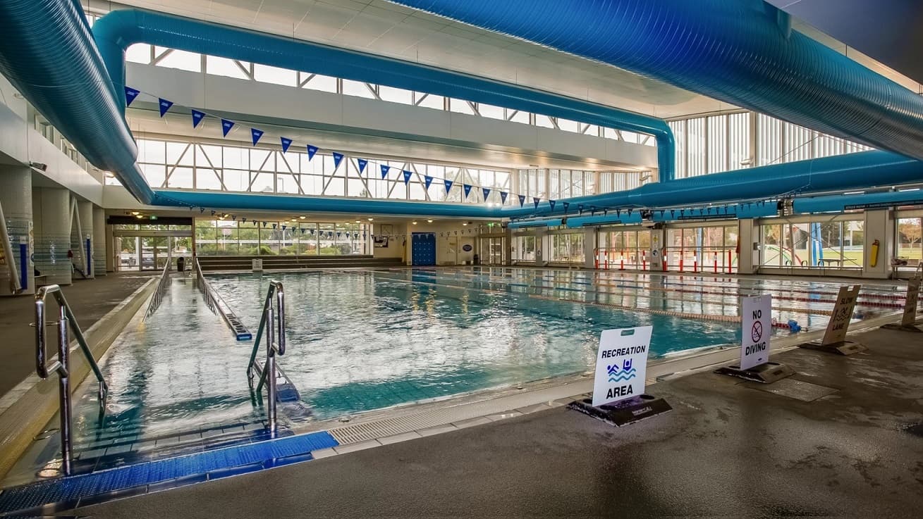 To the left of the pool is the ramp access and at the end of the pool there are large glass windows. To the right of the pool are the swim lanes, separated by coloured lane ropes and there are small blue flags suspended from the ceiling next to the large blue air conditioning pipes that run along the ceiling.
