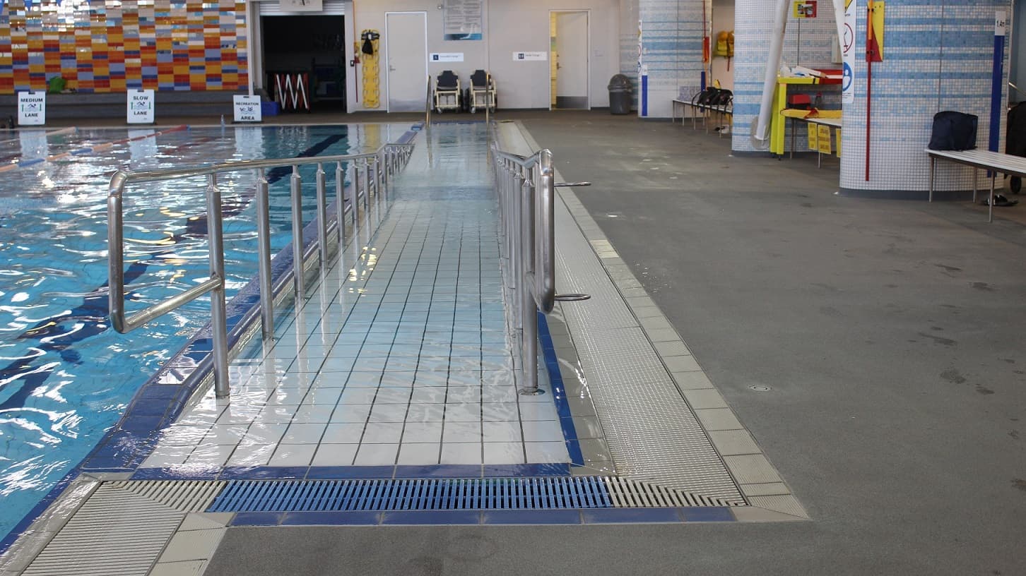 he ramp access to this pool features stainless steel grab rails and is adjacent to the two changing rooms which have two aquatic wheelchairs placed outside. A grey rubbish bin is located to the right of the door in the top right corner. There are three cubicles with blue and white tiling, one with a bench and a bag and the other with yellow and red bags running along the right edge of this space. Signage is positioned along the edge of the pool, advising as to the ‘medium’ and the ‘slow’ lanes.