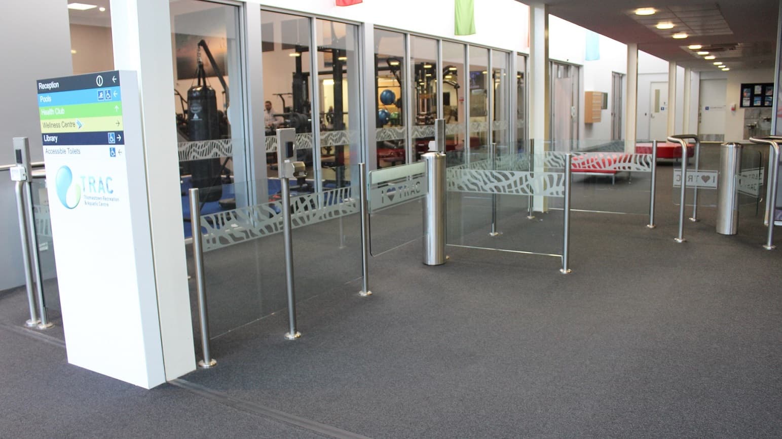A TRAC direction board is set against a pillar by the turnstile gate on the left. The glass-designed wall provides a view of a room with gym equipment. Towards the end of this space is a seating area with two long red seats, close to two white doors. On the wall, there is a brown box on the left, situated next to another small box.