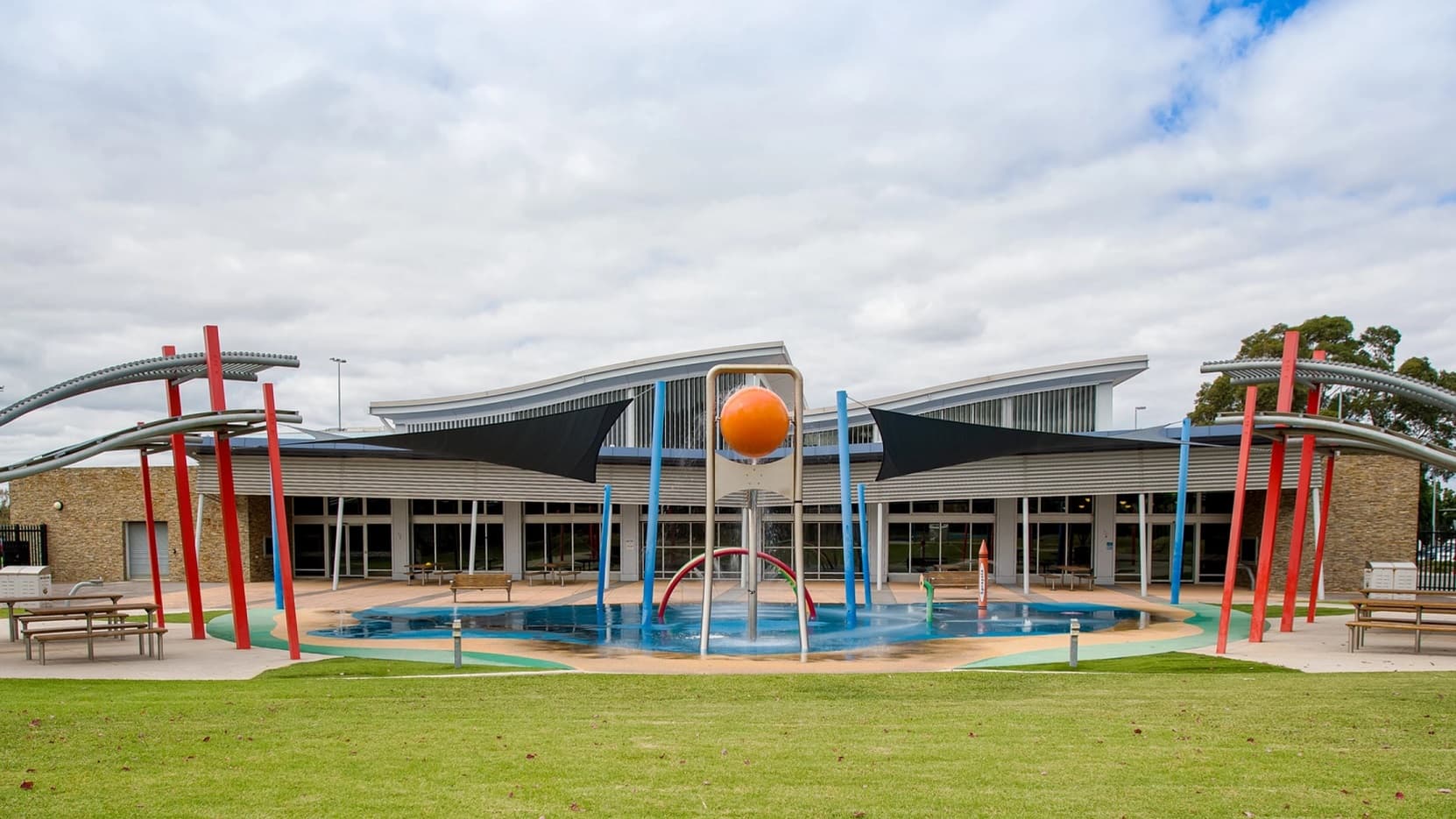 This image is of the outdoor aquatic facility which features two large shaded cloths and has a large orange ball situated in the centre over the small splash pool. There are red poles to the left and to the right and the main building sits in the background. The foreground is green grass and there are seating areas to the left and to the right. Large trees and the sky can be seen in the background.