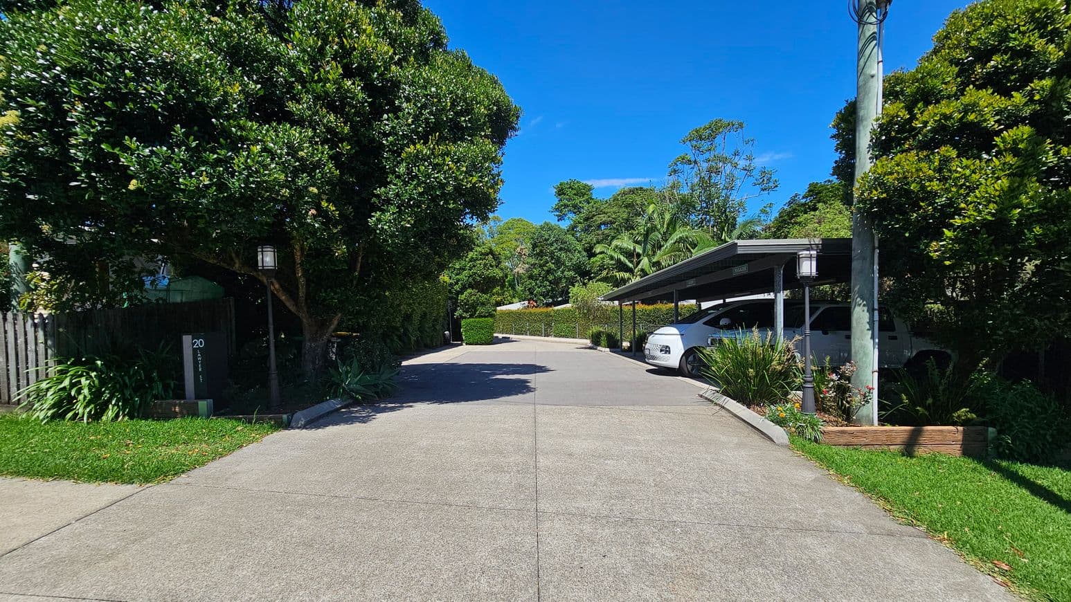 A driveway with a concrete surface leads to a covered parking area on the right, where a white car is parked. On the left, a large tree and a wooden fence are visible, with a house number sign marked "20". The area is surrounded by lush greenery, including bushes and trees. A lamp post stands near the parking area, and a power pole is visible. The scene is well-lit, suggesting a sunny day.