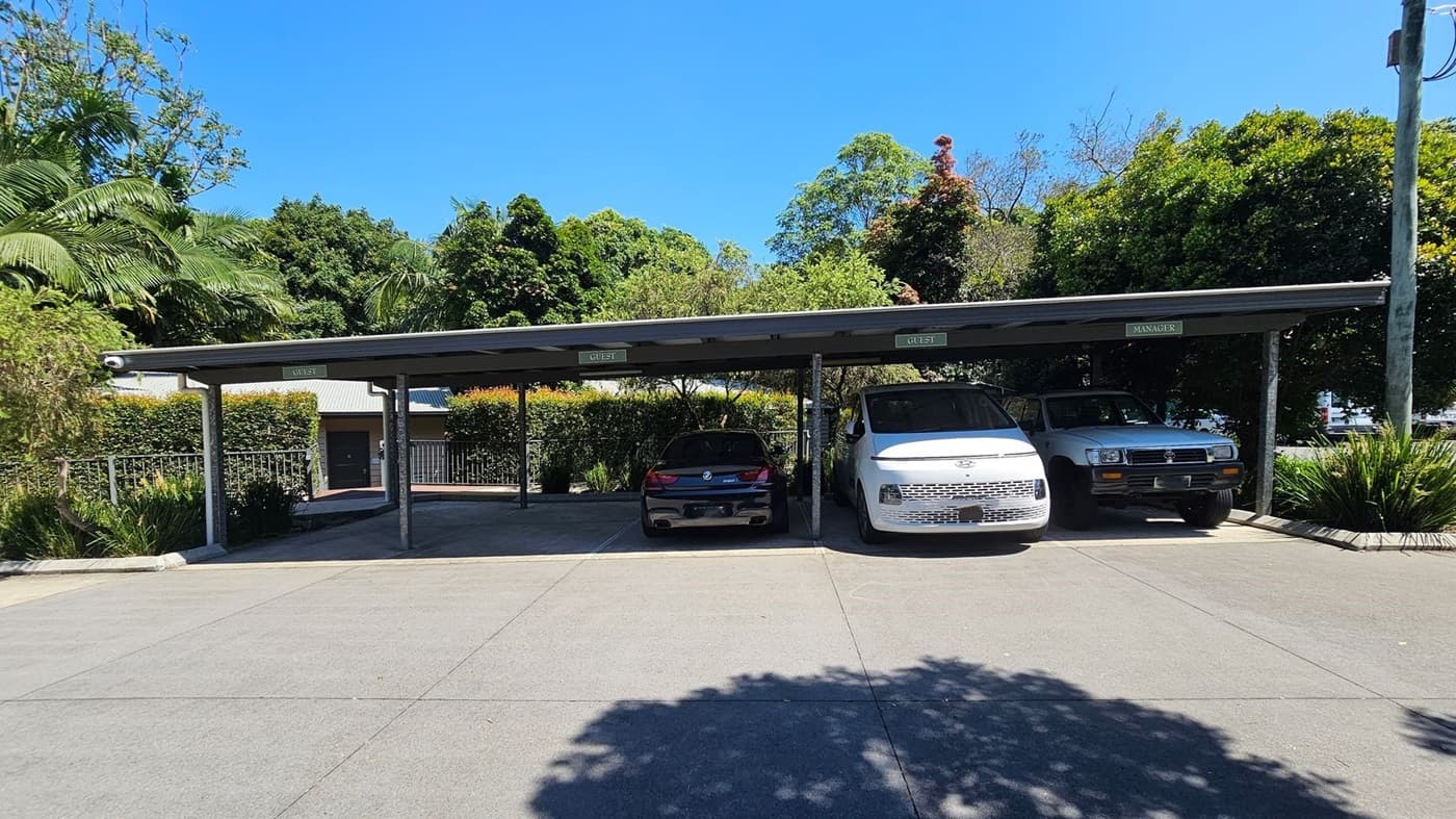 A covered parking area with three vehicles parked side by side. On the left, a black car is parked under a sign marked "GUEST". In the middle, a white van is parked under another "GUEST" sign. On the right, a grey vehicle is parked under a "MANAGER" sign. Behind the parking area, there is a hedge and a building with a metal roof. Lush green trees are visible in the background. The ground is paved with concrete, and there are shadows from the trees.