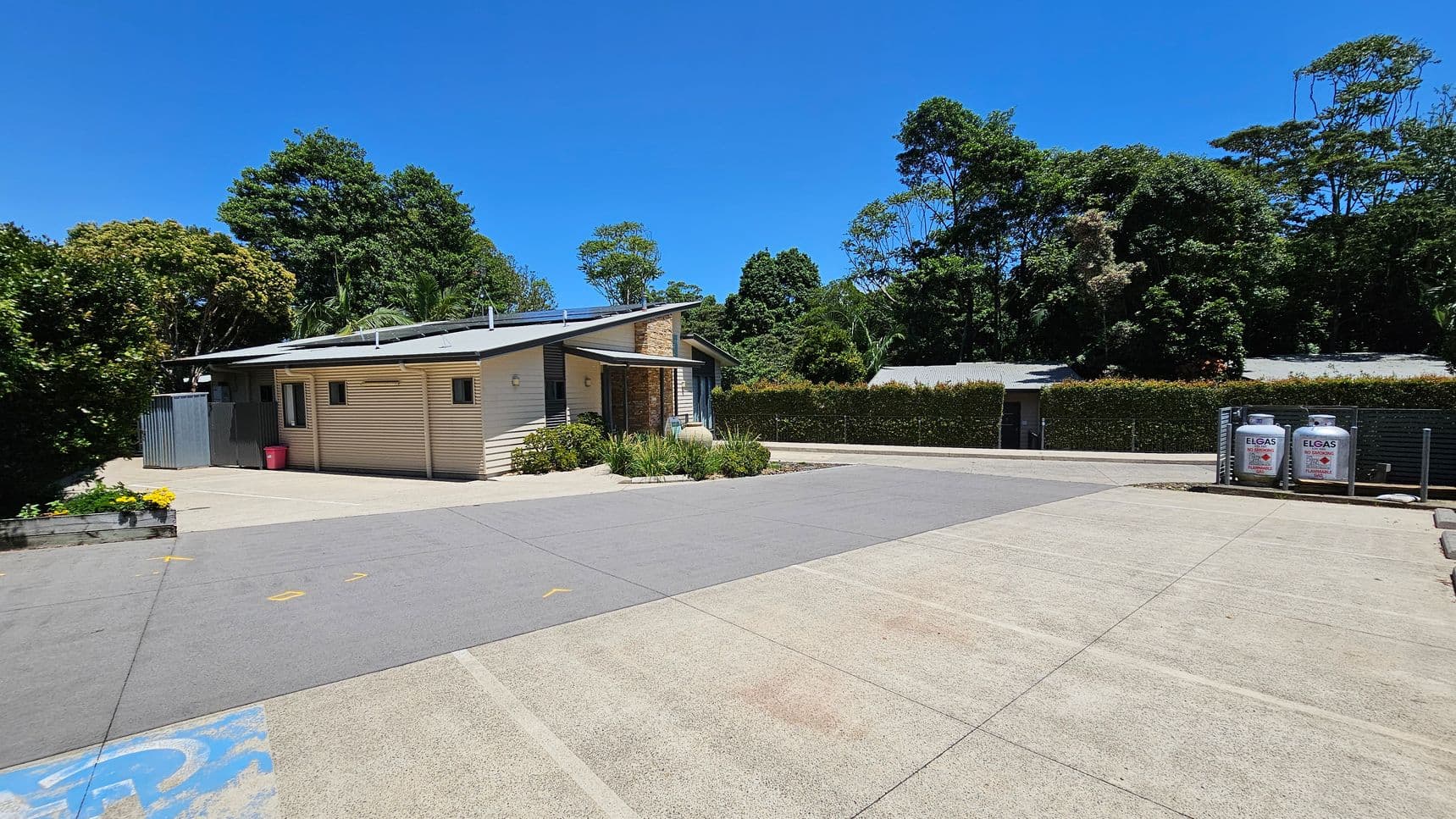 A small, single-storey building with a light-coloured exterior is on the left, surrounded by greenery and trees. In front of the building, there is a paved area with a few yellow markings. On the right, two large gas cylinders are secured in a metal frame. A hedge runs along the background, separating the area from more trees. The ground is a mix of concrete and painted surfaces.