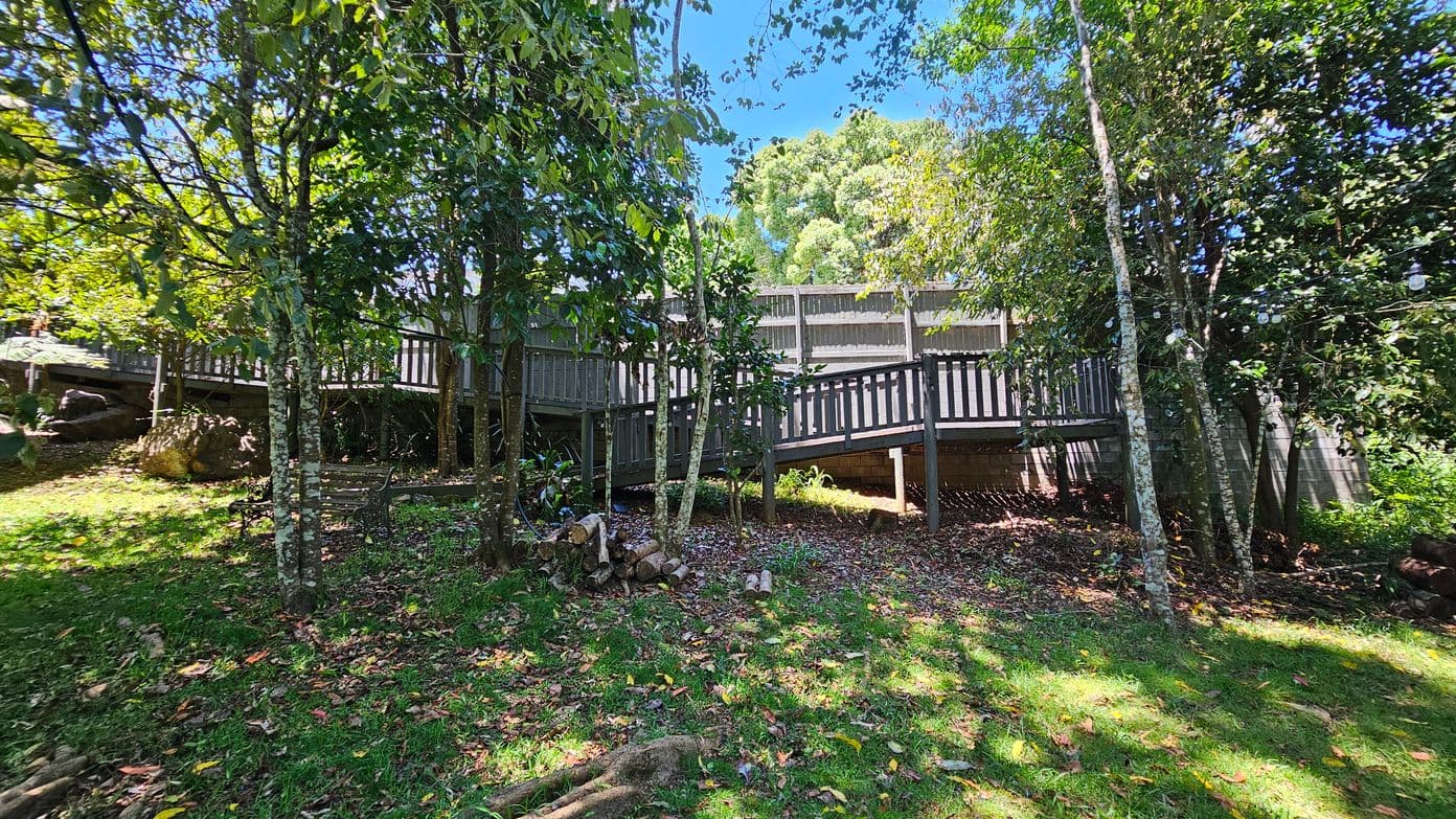 A wooden walkway elevated on stilts runs horizontally across the middle of a lush, green garden. The foreground is covered with grass and scattered leaves, with a few logs piled near the centre. Tall trees with dense foliage surround the area, casting dappled shadows on the ground. In the background, a wooden fence is partially visible through the trees. The scene is brightly lit, suggesting a sunny day.