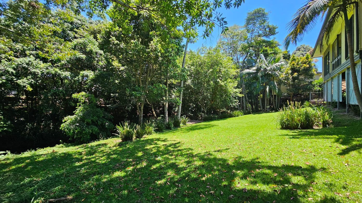 A lush garden with a well-maintained green lawn stretches across the foreground. On the left, dense trees and shrubs create a natural border. To the right, a house with a wooden exterior and a small deck is partially visible, surrounded by tall palm trees. The background is filled with more trees, providing a sense of seclusion and tranquillity. Shadows from the trees dapple the grass, adding texture to the scene.