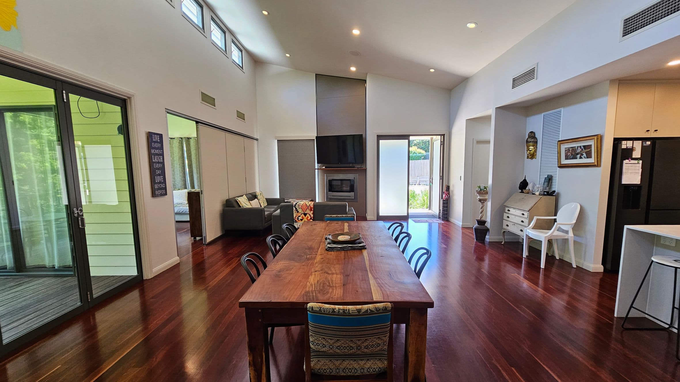 A spacious living area with polished wooden floors. On the left, large glass sliding doors lead to an outdoor area. A grey sofa with cushions is visible in the middle, near a wall-mounted TV above a fireplace. A long wooden dining table with chairs is centrally placed. On the right, a white chair, a small desk, and a black refrigerator are visible. The ceiling is high with recessed lighting, and there are small windows near the top. A door at the back opens to a garden.