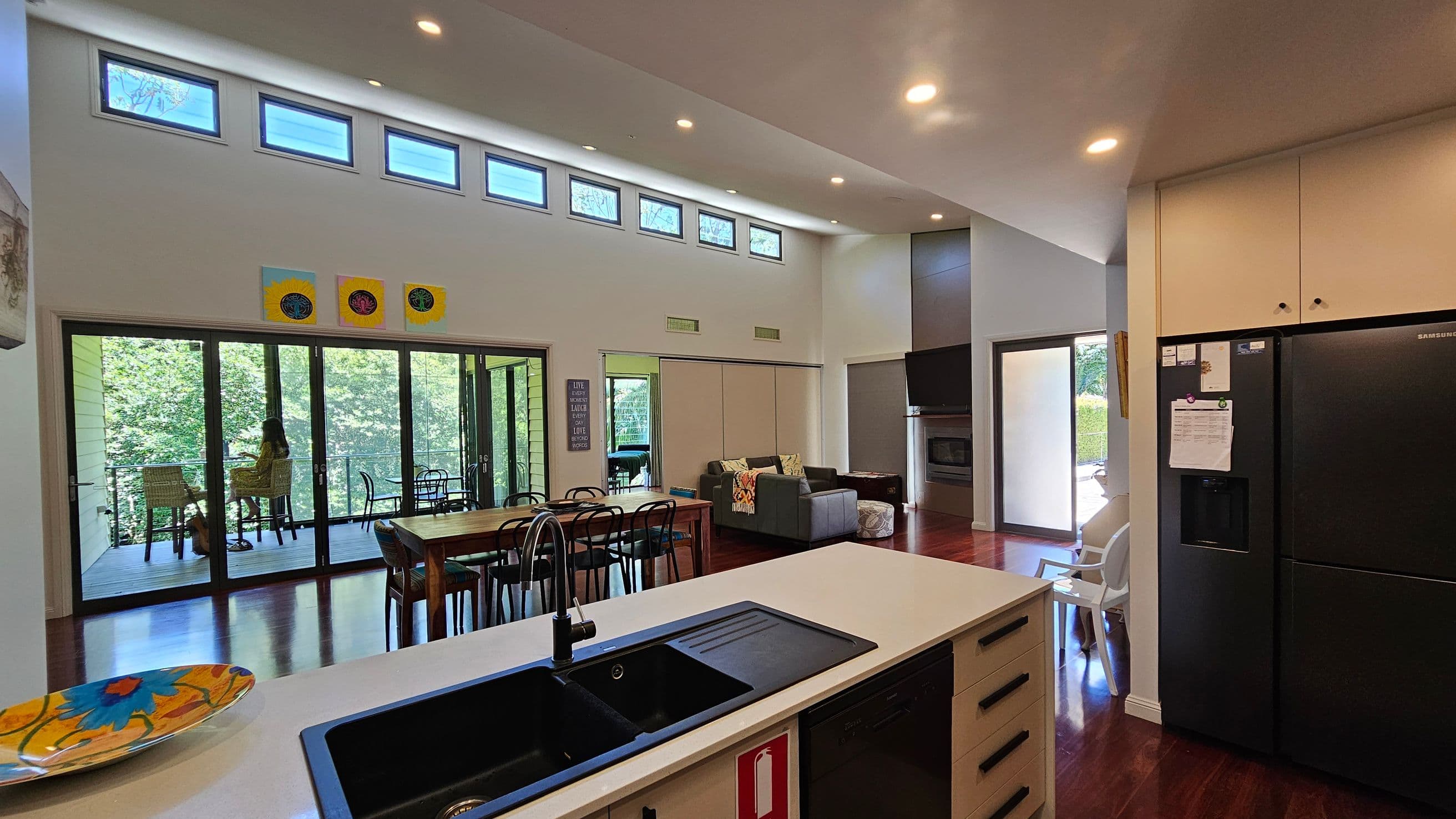 A modern open-plan living space with a kitchen in the foreground featuring a black sink and a colourful plate on the counter. To the right, a black refrigerator stands against a wall with white cabinets. In the middle, a dining table with chairs and a grey sofa are visible. Large glass doors on the left open to a balcony with a person sitting at a table. The room has high ceilings with small windows near the top and wooden flooring. Bright artwork is displayed above the doors.