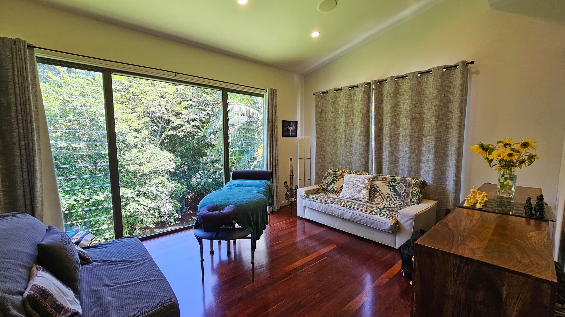 A room with wooden flooring and a large window on the left, offering a view of lush green trees. In front of the window is a massage table with a teal cover. To the right, a white sofa with patterned cushions is placed against a wall with grey curtains. A wooden cabinet on the far right holds a vase of sunflowers and a chessboard. The ceiling is light-coloured with recessed lighting.
