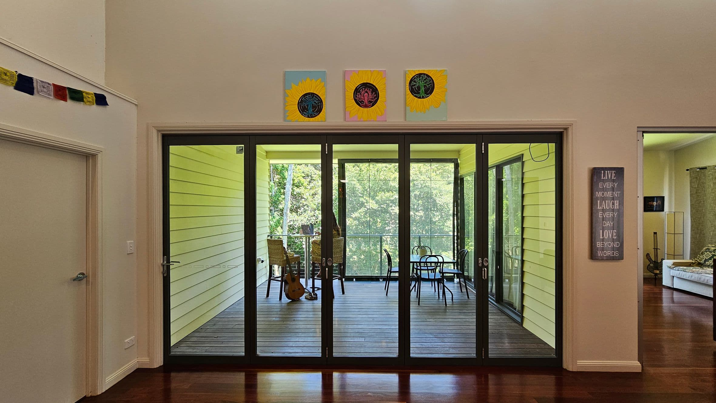 Glass doors in the centre open to a wooden-floored patio with wicker chairs and a guitar. The background shows lush greenery. Above the doors are three sunflower paintings. To the left, a string of colourful flags hangs near a closed door. On the right, a wall sign reads "LIVE EVERY MOMENT LAUGH EVERY DAY LOVE BEYOND WORDS" next to an open doorway leading to a room with a sofa and a guitar. The floor is dark wood.