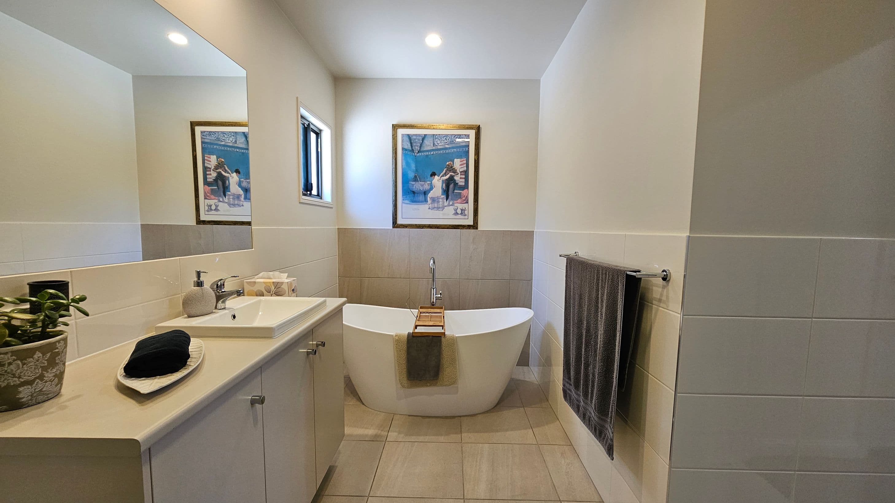 A bathroom with a white freestanding bathtub in the centre, featuring a wooden bath tray and a towel draped over the side. Above the tub is a framed picture. To the left, a countertop with a sink, soap dispenser, tissue box, and a potted plant. A large mirror is mounted on the wall above the sink. On the right, a towel hangs on a rail. The walls are tiled halfway up, and the floor is covered with light-coloured tiles. A small window is above the bathtub.