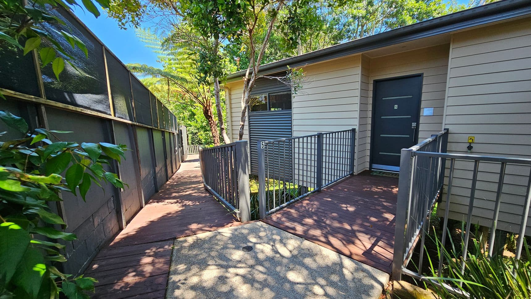 A pathway leads to a building entrance with a dark door marked with the number 5. The building has light-coloured horizontal siding. On the left, a tall fence with a mesh top runs alongside the path, partially shaded by trees. The path is made of wooden planks, and there are metal railings on both sides near the entrance. Lush greenery surrounds the area, with trees and bushes providing shade. The ground is a mix of concrete and wood, with dappled sunlight filtering through the leaves.