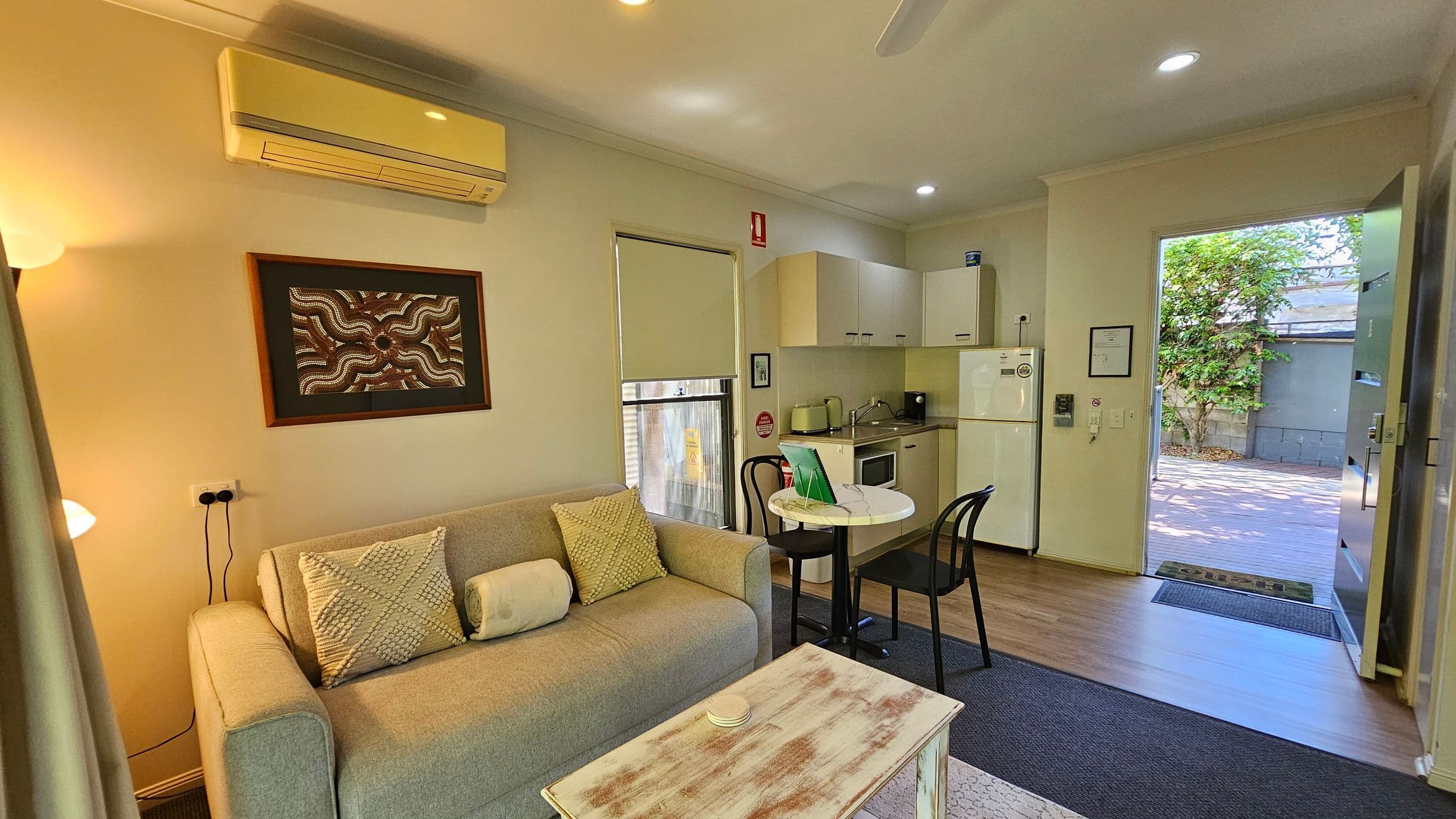A cosy living area with a light grey sofa adorned with patterned cushions on the left. Above the sofa is a framed artwork and an air conditioning unit. A small wooden coffee table sits in front of the sofa. To the right, there's a compact kitchen with white cabinets, a fridge, and a small round dining table with two chairs. The floor is a mix of carpet and wood. An open door leads to an outdoor area with greenery visible. The ceiling has recessed lighting.