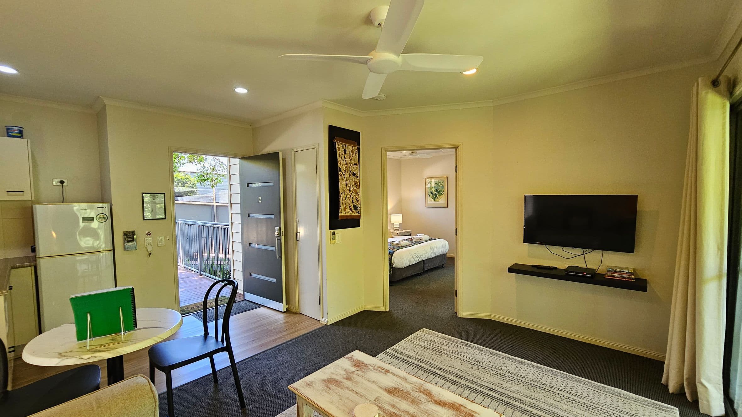 A compact living space with a ceiling fan. On the left, there's a small kitchen area with a fridge and a round table with two chairs. The entrance door is open, leading to a wooden deck outside. In the centre, a doorway reveals a bedroom with a bed and bedside lamp. On the right, a wall-mounted TV is above a shelf with a few items. The floor is a mix of carpet and wood, and curtains are partially visible on the right.