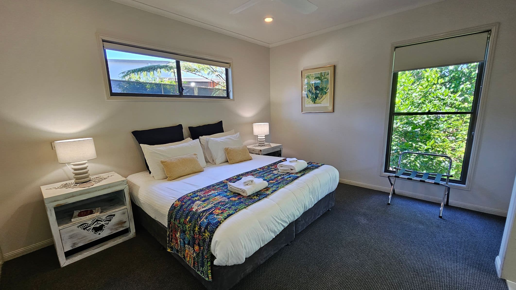 A bedroom with a large bed in the centre, adorned with white linens, two beige cushions, and a colourful throw. Two neatly folded towels are placed on the throw. On either side of the bed are white bedside tables with lamps. A small window above the bed and a larger window on the right let in natural light, with greenery visible outside. A luggage rack is positioned near the larger window. The floor is carpeted in a dark grey tone, and a framed picture hangs on the wall.